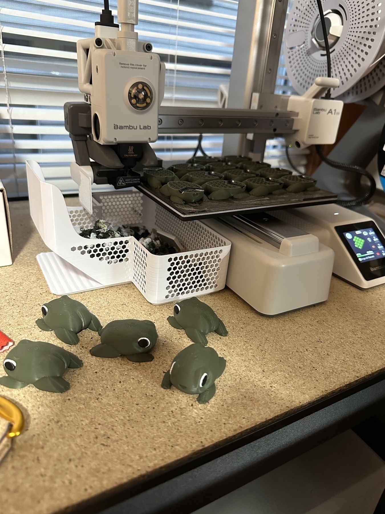 Bamboo lab A1, mini 3-D printer in progress printing a full bed of frogs. On the table in front of the printer are five completed frogs they have a little dump truck booties.